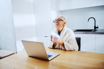 Sick attractive caucasian blond senior woman covered with blanket sitting at dining table and holding mug with tea and handkerchief. On table is laptop.