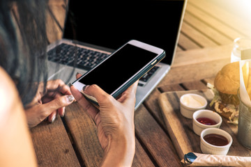 Woman using a smart phone with laptop at the restaurant. Hamburger and french fries on the table with light on the right. 