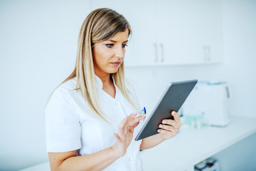 Portrait of beautiful blond female lab assistant in white uniform using tablet while standing in laboratory.