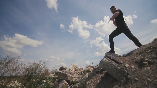 Handsome, young man with a beard in a black T-shirt demonstrates mastery with nunchaku against the sky background.