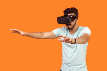 Portrait of attentive gamer, brunette man with beard in t-shirt wearing vr glasses, stretching arms forward while playing virtual reality game, 3d headset. studio shot isolated on orange background