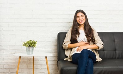 Young arab woman sitting on the sofa laughs happily and has fun keeping hands on stomach.