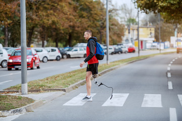 Full length of handsome caucasian handicapped sportsman in sportswear, with artificial leg and backpack crossing street.