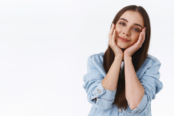 Tenderness, beauty and emotions concept. Cute and gentle feminine young 20s woman in denim jacket, tilt head, touching cheeks and smiling dreamy, look with admiration and affection, white background