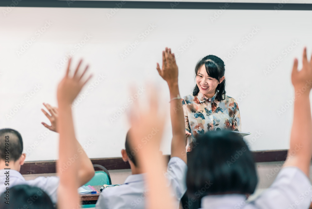 Wall mural An smiling Asian female high school teacher teaches the white uniform students in the classroom by asking questions and then the students raise their hands for answers.