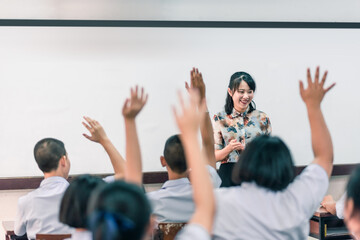 An smiling Asian female high school teacher teaches the white uniform students in the classroom by asking questions and then the students raise their hands for answers.