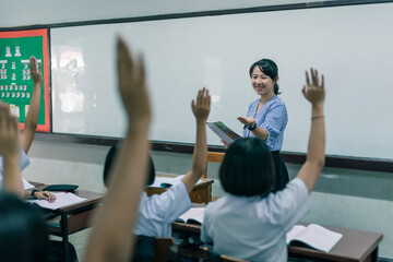 Fototapeta na wymiar An smiling Asian female high school teacher teaches the white uniform students in the classroom by asking questions and then the students raise their hands for answers.