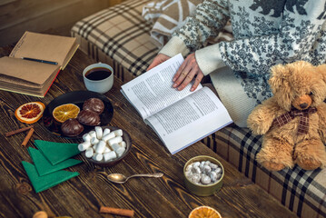 Woman in a sweater reading a book with a mug of coffee in the evening in a warm Christmas atmosphere. Cozy new year mood