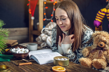 Young girl in a sweater reading a book with a mug of tea in the evening in a warm Christmas...