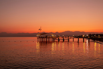 Beautiful golden sunset sea landscape with wooden pier of hotel resort, shiny sea water with reflected lights on surface and silhouettes of dark mountains at horizon. Horizontal color photography.