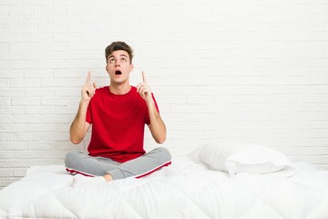 Young teenager student man on the bed pointing upside with opened mouth.
