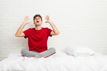 Young teenager student man on the bed screaming to the sky, looking up, frustrated.