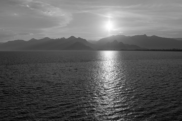 Aerial top view of peaceful calm dark blue sunny sea water surface, cloudy sunset or sunrise sky and silhouettes of mountains with soft sun over peaks. Black and white horizontal color photography.