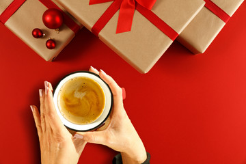 Christmas composition of ecological gifts with red ribbon and bow.Female hands wrapped gifts.Cup of coffee in women's hands and time of Christmas rest.Top view,flat lay,copy space,aerial xmas photo.