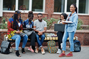 Fototapeta premium Group of five african college students spending time together on campus at university yard. Black afro friends studying at bench with school items, laptops notebooks.
