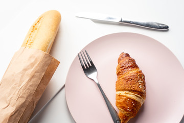 Fresh baguette and croissant, a knife and a fork on  the white table in the kithchen in the morning. Top view