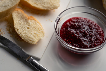 A glass bowl with sweet raspberry jam, slices of baguette and a knife on a white table in the kitchen in the morning. 