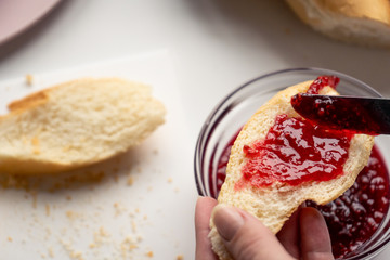 Woman spreads a lot of sweet red raspberry jam on a slice of baguette for breakfast in a kitchen in the morning. Close-up