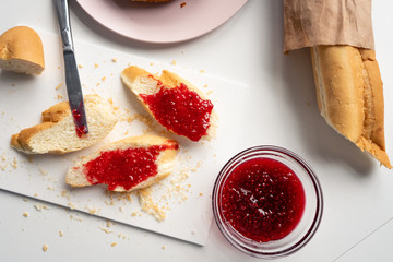 Fresh sliced ​​baguette with sweet raspberry jam for vitamin breakfast and a round plate on a white table in the kitchen. Top view