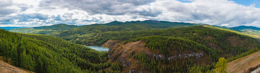 Naklejka na ściany i meble Panorama of mountain lake on background of beautiful snowy mountain peaks in cloudy weather. Mountain lake in the forest. View of lake on the background of blue sky