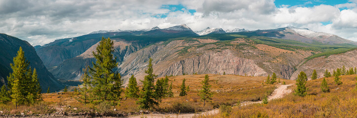 Panorama of road to the gorge of mountains on background of beautiful snowy mountain peaks in cloudy weather. Mountain road in the forest. View of trail on the background of blue sky