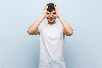 Young caucasian man wearing a white tshirt showing okay sign over eyes