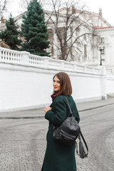 Fototapeta premium Beautiful young stylish brunette woman wearing midi green coat, black backpack, purple scarf walking through the city street. Trendy casual outfit. Street fashion. Everyday winter, autumn, spring look