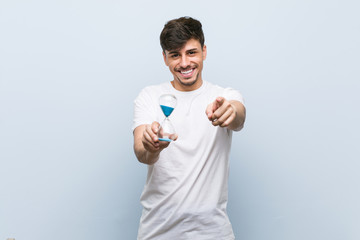 Young hispanic man holding a hourglass cheerful smiles pointing to front.