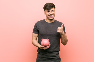 Young hispanic man holding piggy bank smiling and raising thumb up