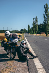 Obraz premium A male biker is sitting by the motorcycle on the side of the road. Dressed in motorcycle equipment. The concept of a long trip on a tourist bike. asphalt road, vertical photo