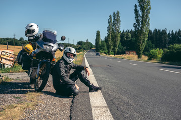 Obraz premium A male biker is sitting by the motorcycle on the side of the road. Dressed in motorcycle equipment. The concept of a long trip on a tourist bike. asphalt road, place for text.