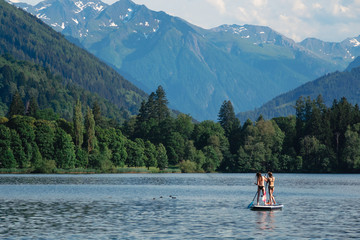 Fototapeta na wymiar Two young beautiful girl-surfer riding on the stand-up SUP board in the clear waters of the Alpine mountains on the background. Lake 