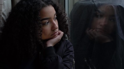 Sad depressed beautiful female mixed race African American girl teenager young woman teen wearing a black jacket sitting looking out of a window - Powered by Adobe
