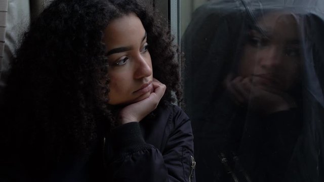 Sad depressed beautiful female mixed race African American girl teenager young woman teen wearing a black jacket sitting looking out of a window 