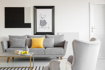 Big comfortable wing back armchair next to long grey scandinavian sofa with pillows in bright living room interior