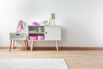 Small toy in pink clothes on white wooden nightstand table next to white cabinet with in elegant interior
