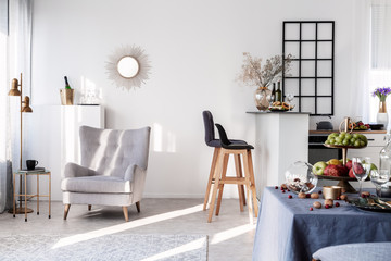 Trendy grey armchair next to two black wooden bar stools in fashionable kitchen and dining room...