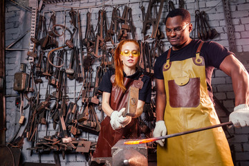couple in love redhaired ginger young european woman and afro-american man wearing leather apron working blacksmith workshop.small family international business concept
