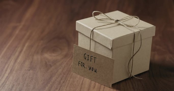 Slow motion man hand put gift for you card next to brown paper gift box on walnut table