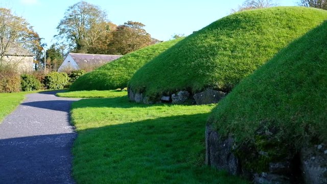 handheld wide shot of the archeological enclosure of Knowth, Boyne Valley Tombs with green grass covered tombs and an ancient stone cottage in the background on a sunny blue sky day and no people.
