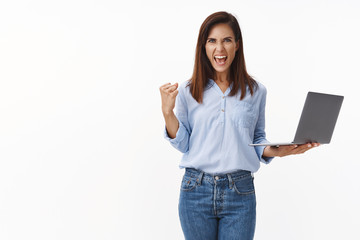 Empowered good-looking businesswoman feel success and joy, hold laptop, fist pump celebrating good deal, shout yeah yes pleased, triumphing excellent news, working hard and motivated