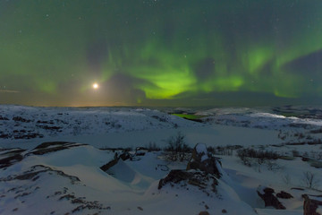 Fototapeta na wymiar Northern lights, aurora in the sky at night. Hills and rocks covered with snow. Moon in the sky.