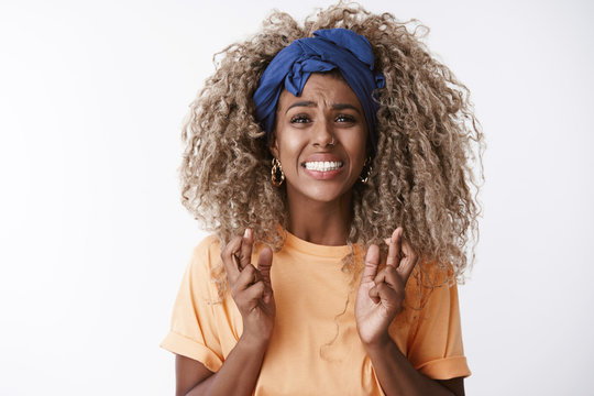 Close-up worried and anxious, troubled young hopeless african-american curly blond woman in orange t-shirt, headband, clench teeth desperate look camera, cross fingers good luck, praying