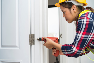 Young Asian carpenter woman, technician or worker using power drill bit installing screwed a door hinges or hinges on wooden door. House, Renovation or Drilling Equipment Construction service concept