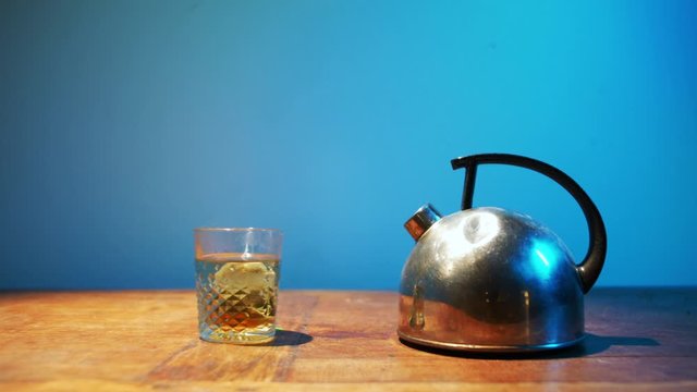 Push-in on tea in glass and metal kettle on wooden table in front of blue wall