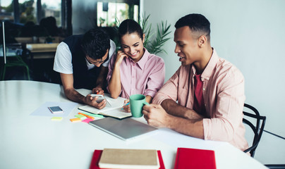 Successful male and female colleagues smiling during brainstorming meeting for creating ideas for startup project, positive multicultural group of friends talking near desktop using cellphone
