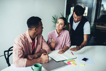 Intelligent male and female colleagues cooperating on information for startup project sitting at desktop in coworking space, smart group of diverse students preparing course work presentation