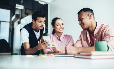Multicultural male and female colleagues smiling during discussion about content text in social networks, successful men and woman sitting at desktop and feeling good from time for collaboration