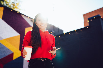 Cheerful hipster girl connecting smartphone to 4G internet in roaming, smiling chinese woman in trendy wear using mobile phone for chatting outdoors satisfied with technology communication