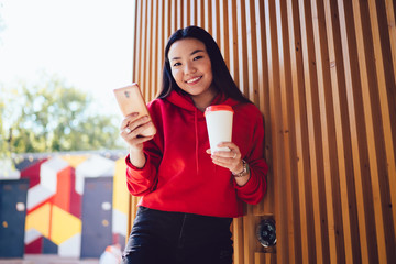 Half length portrait of cheerful female student dressed in stylish red apparel holding takeaway cup and smartphone device for blogging, happy japanese woman enjoying life in technology generation